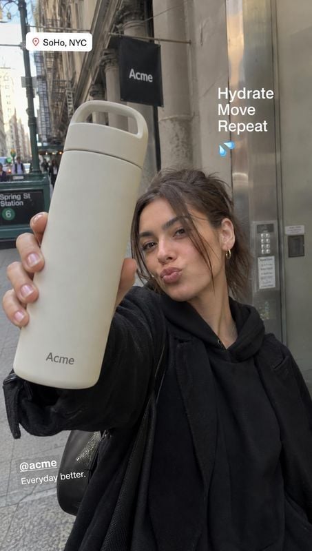 reusable water bottle product in hand product photo: A woman in a black hoodie holds a cream reusable bottle toward the camera on a city street, with overlaid…