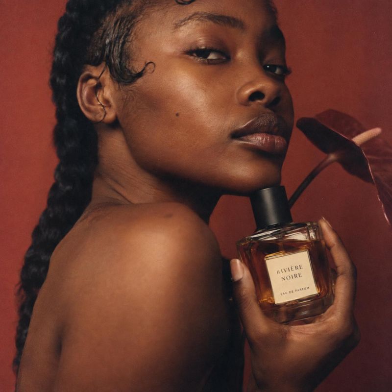 perfume beauty close-up product photo: A woman with sleek braided hair holds an amber perfume bottle against a deep terracotta backdrop, lit warmly for an inti…