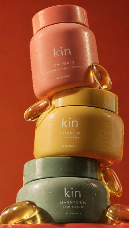 Kin supplement bottles group lineup product photo: Three pastel supplement bottles are stacked vertically with golden softgels arranged around them against a w…