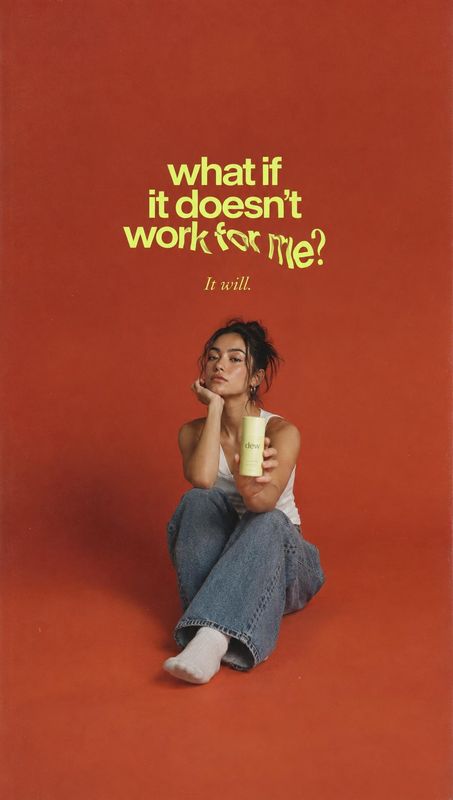 skincare bottle product in hand product photo: A woman in a white tank top and relaxed jeans holds a pale bottle against a vivid rust-red studio backdrop, with…