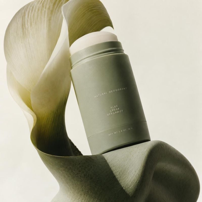 natural deodorant personal care product photo: A sage-green natural deodorant tube rests among sculptural pale-green leaves against a softly lit cream backgrou…