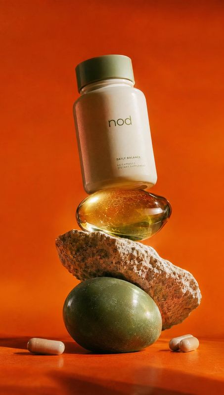 daily balance supplement bottle personal care product photo: A pale supplement bottle and amber glass vessel are balanced with textured stones and capsules aga…