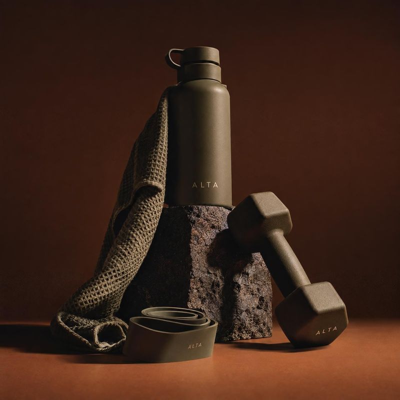 fitness water bottle sports product photo: A matte bottle, dumbbell, rolled bands and textured towel are arranged on a dark rust-toned studio set, lit with dra…