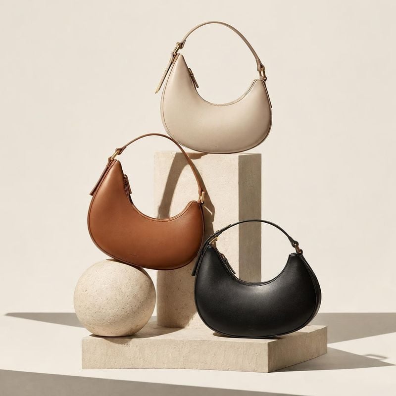 handbags group lineup product photo: Three crescent-shaped handbags in cream, brown, and black are arranged on sculptural stone blocks against a warm beige stu…