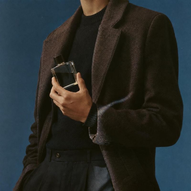 perfume bottle product in hand product photo: A cropped male model in dark, tailored layers holds a clear perfume bottle against a deep blue studio backdrop, w…