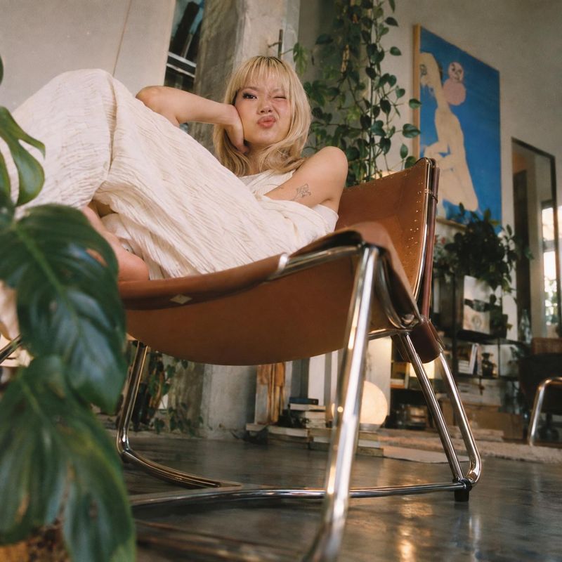 lounge chair lifestyle scene product photo: A woman in a flowing white outfit lounges in a brown leather sling chair amid leafy plants and art, in a warm, rela…