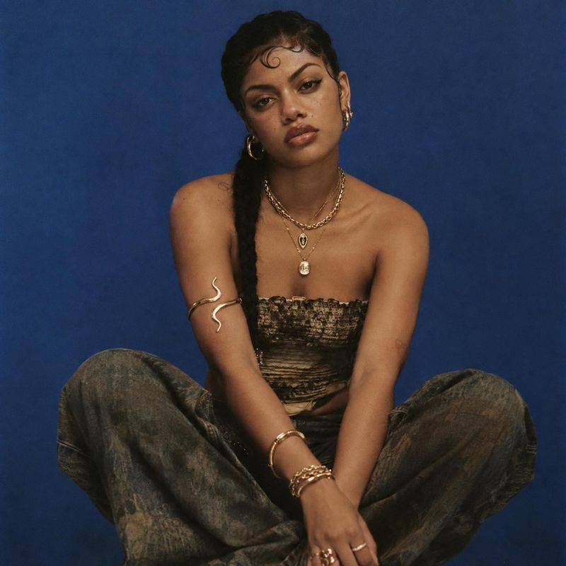 gold jewelry on-body detail product photo: A model poses against a deep blue studio backdrop in a patterned strapless top and relaxed trousers, styled with lay…