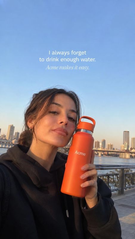 reusable water bottle lifestyle scene product photo: A woman holds an orange reusable bottle close to her face on a waterfront promenade, with the city skyline…