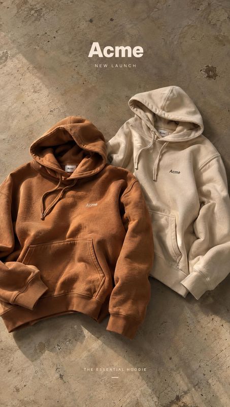 hoodies flat lay product photo: Two hoodies in rust brown and cream are arranged on a textured concrete surface beneath prominent launch typography, with warm…