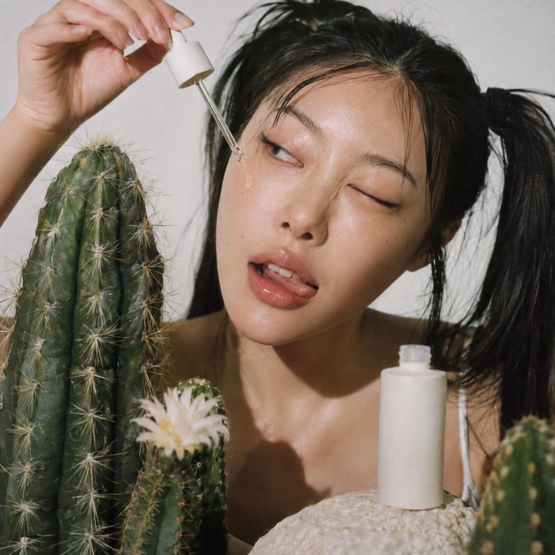 skincare serum beauty close-up product photo: A close beauty portrait shows a woman applying serum with a dropper among cacti and pale stone, lit with soft nat…