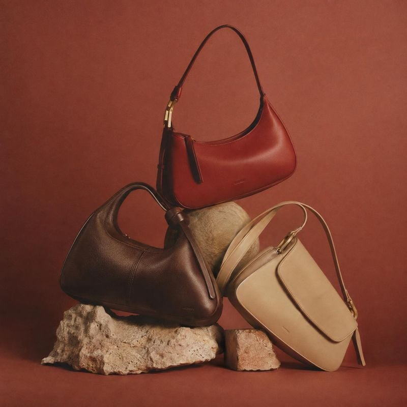 handbags group lineup product photo: Three leather handbags in red, dark brown, and beige are artfully arranged on rough stone props against a warm terracotta…