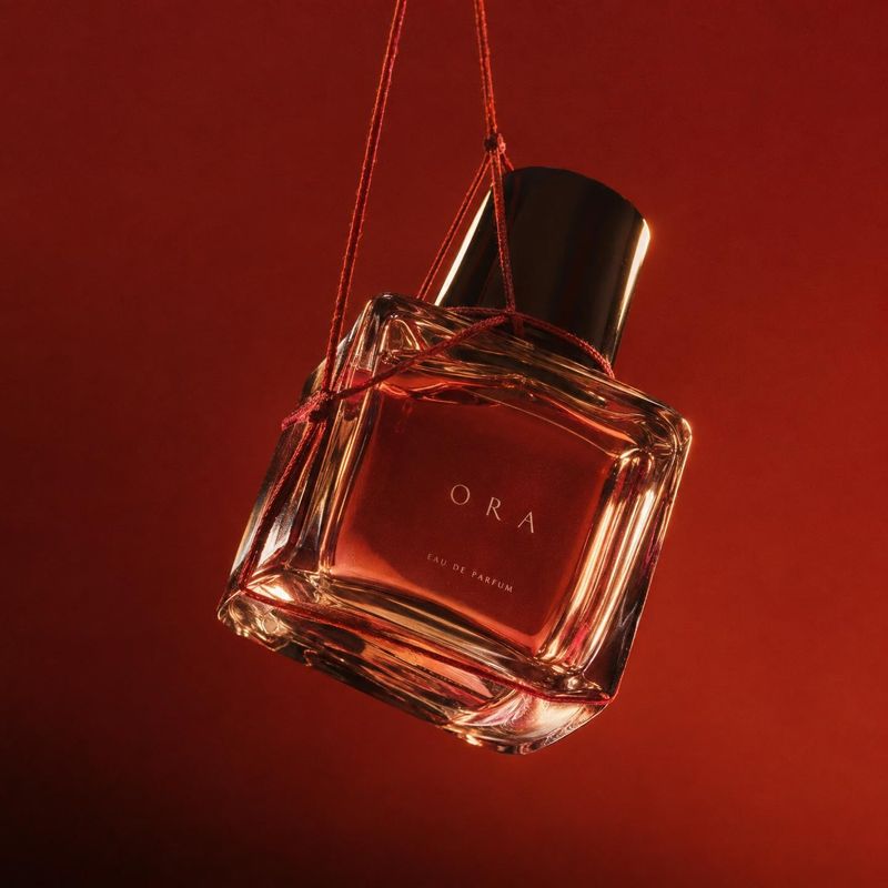 perfume bottle floating product product photo: A glass perfume bottle hangs from red cords against a deep red studio backdrop, lit with dramatic warm highlight…
