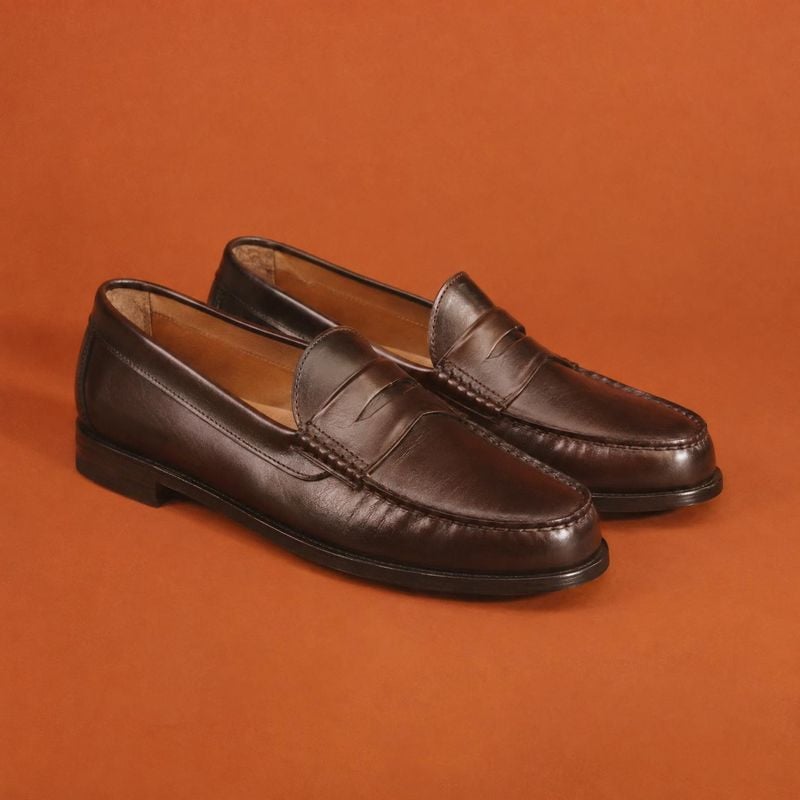brown leather loafers fashion product photo: A pair of polished dark brown leather loafers is arranged at an angle on a seamless burnt-orange studio backdrop,…