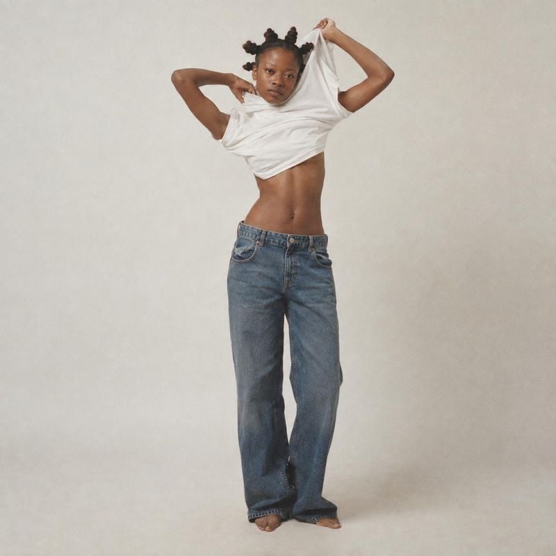 jeans action & motion product photo: A model in loose blue jeans lifts a white shirt in a softly lit neutral studio, creating a relaxed, playful fashion image.
