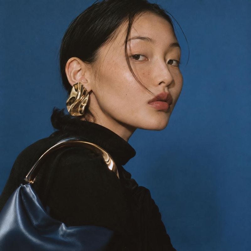 gold statement earrings on-body detail product photo: A close fashion portrait shows a woman in a black outfit wearing sculptural gold earrings and carrying a…