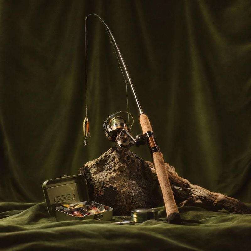 fishing rod and tackle gear sports product photo: A fishing rod, reel, lure, and open tackle box are artfully arranged on rock and draped fabric, with dramatic…