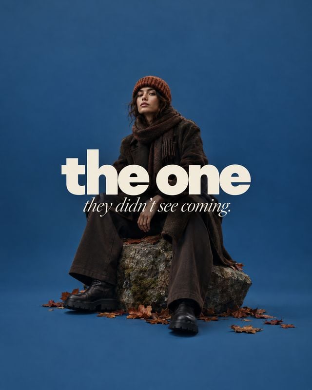 winter outfit autumn product photo: A model in a dark, layered winter outfit and knit beanie sits on a rock amid fallen leaves against a deep blue studio backd…