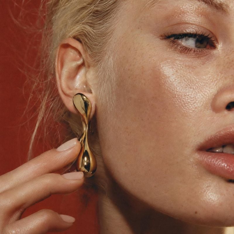 gold drop earring beauty close-up product photo: A close-up portrait shows a woman holding a sculptural gold drop earring to her ear against a warm red studio…