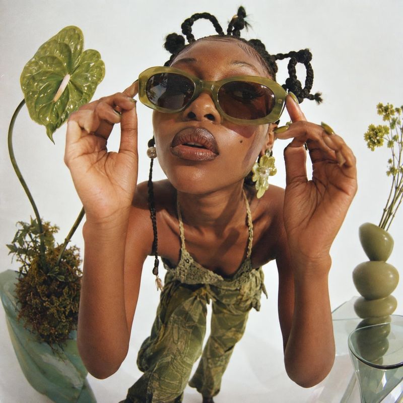 sunglasses on-body detail product photo: A model in sculptural braids adjusts oversized olive-toned sunglasses in a bright studio, surrounded by leafy plants a…