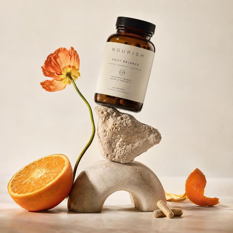 daily balance supplement bottle personal care product photo: An amber supplement bottle balances on sculptural stone beside an orange, a vivid flower, and caps…