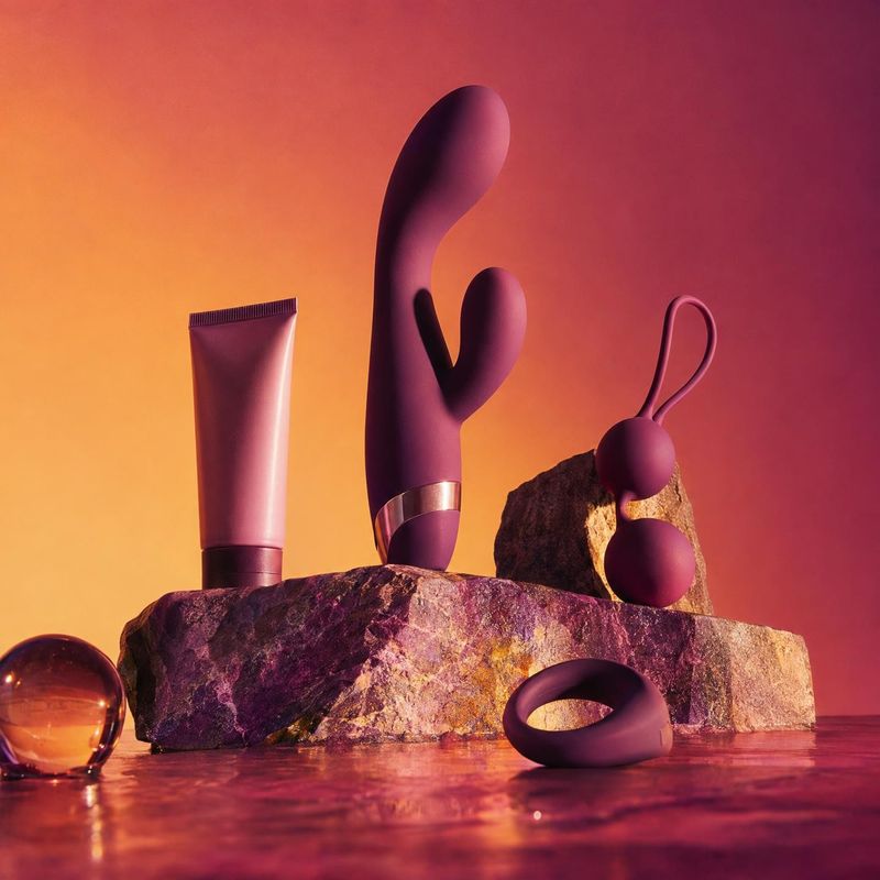 intimate wellness products personal care product photo: A tube and several sculptural purple intimate wellness devices are arranged on rugged stone against a g…