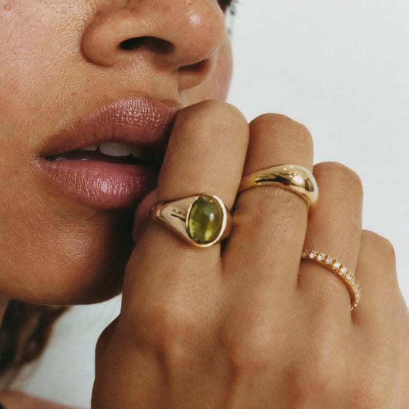 gold rings close-up detail product photo: A close crop shows a model wearing sculptural gold rings and a green gemstone ring near her lips, with soft neutral l…