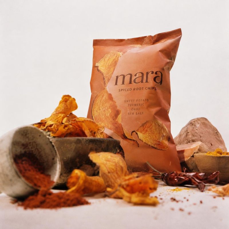 spiced root chips ingredients product photo: A copper-colored bag of spiced root chips stands among chips, sweet potatoes, spices and rustic bowls on a pale st…