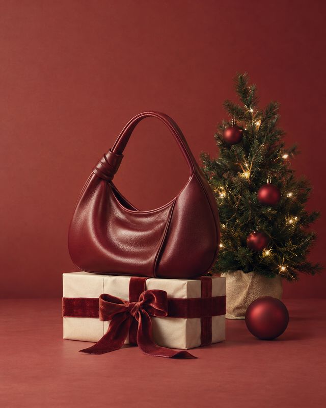 burgundy handbag christmas product photo: A glossy burgundy handbag sits on a ribbon-wrapped gift beside a softly lit Christmas tree and ornaments, styled agai…