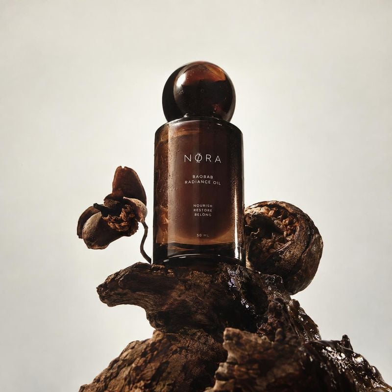 baobab radiance oil ingredients product photo: An amber glass oil bottle stands among dark, sculptural botanical elements against a softly lit pale studio back…