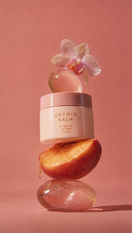 orchid balm moisturizer ingredients product photo: A pale pink orchid balm jar is balanced with a glossy peach, translucent stones, and an orchid against a war…