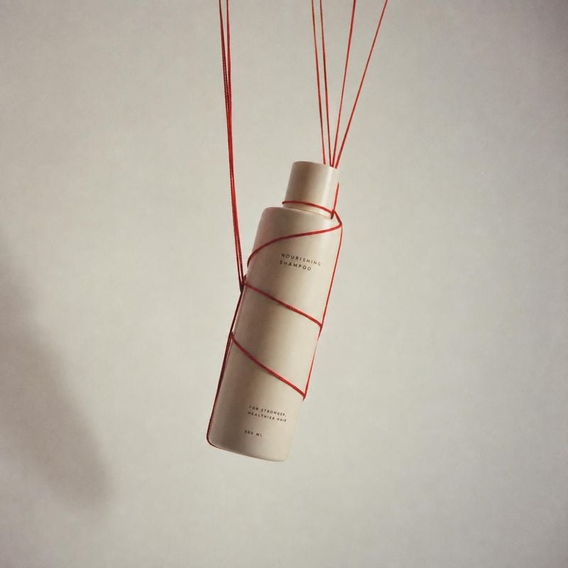 nourishing shampoo beauty product photo: A minimalist shampoo bottle hangs suspended by fine red cords against a softly lit warm-white studio backdrop, creatin…