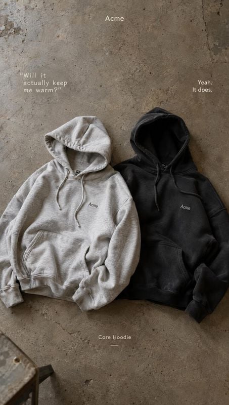 hoodies flat lay product photo: Two hoodies in light gray and black lie side by side on a rough concrete floor, with small white campaign text and a muted, und…