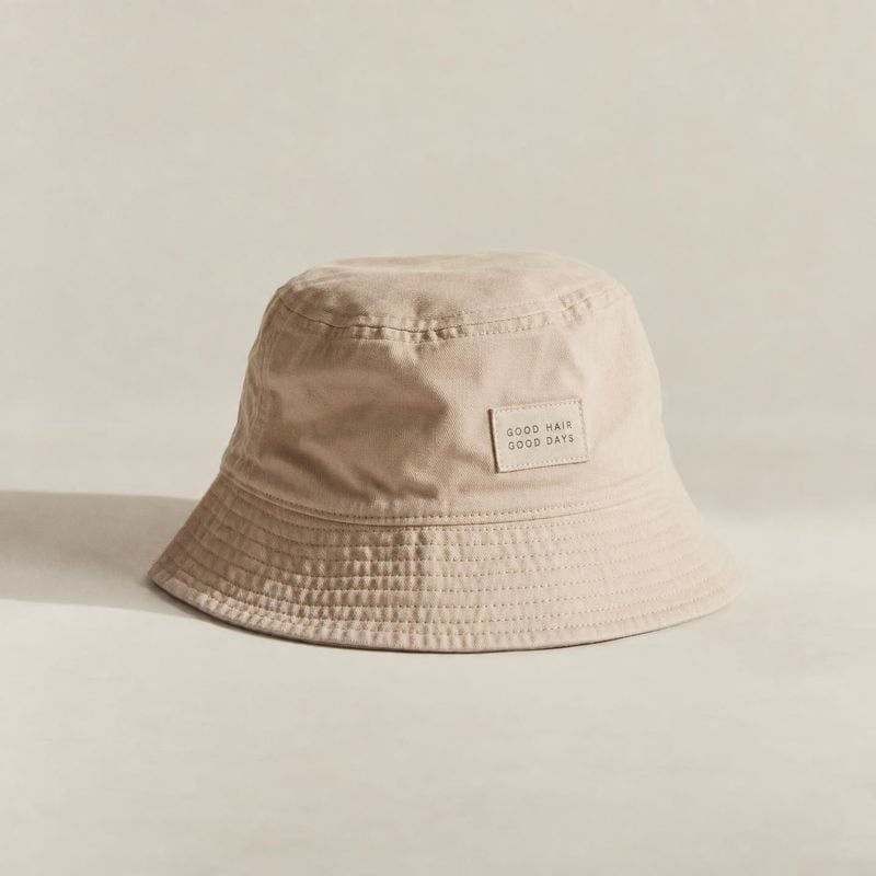 beige bucket hat fashion product photo: A beige bucket hat with a small stitched label sits on a pale studio surface, softly lit with gentle shadows and ample…