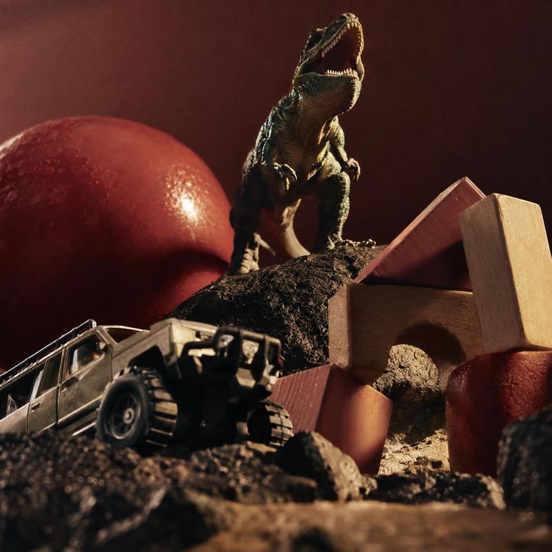 dinosaur and off-road vehicle toy set lifestyle scene product photo: A roaring dinosaur figure towers over a toy off-road vehicle and wooden blocks in a rugged…