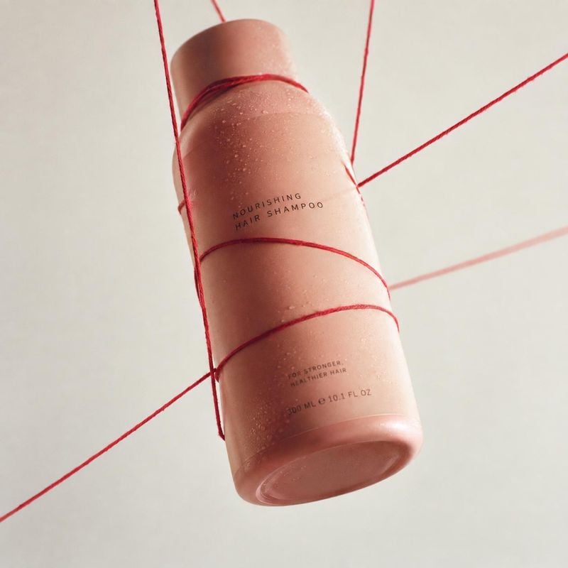 shampoo bottle beauty product photo: A peach-toned shampoo bottle wrapped in fine red cord hangs against a pale studio backdrop, with soft warm lighting and a…