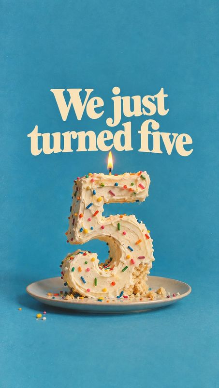 number-five birthday cake food and drink product photo: A sprinkle-covered number-five cake with a lit candle sits on a plate against a bright blue studio back…