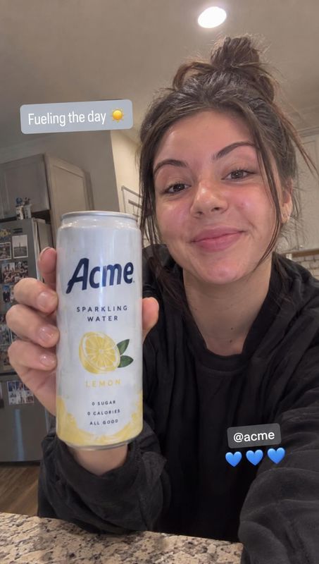lemon sparkling water product in hand product photo: A woman in a dark sweatshirt smiles and holds a lemon sparkling water can toward the camera in a home kitc…