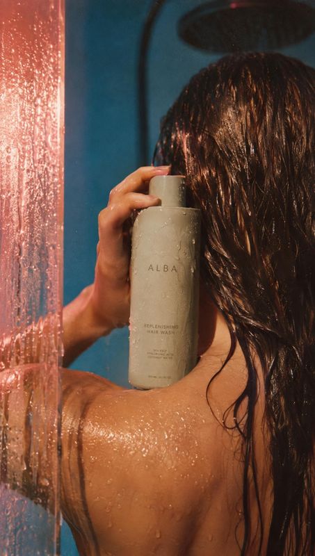 hair wash product in hand product photo: A woman with wet, dark hair holds a neutral-toned hair wash bottle in a shower, with water beading on her skin and war…