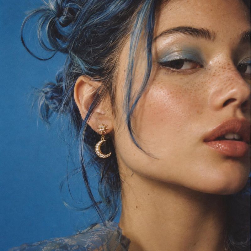 crescent moon drop earring beauty close-up product photo: A close beauty portrait shows a woman with blue-toned eye makeup and a gold crescent moon earring aga…