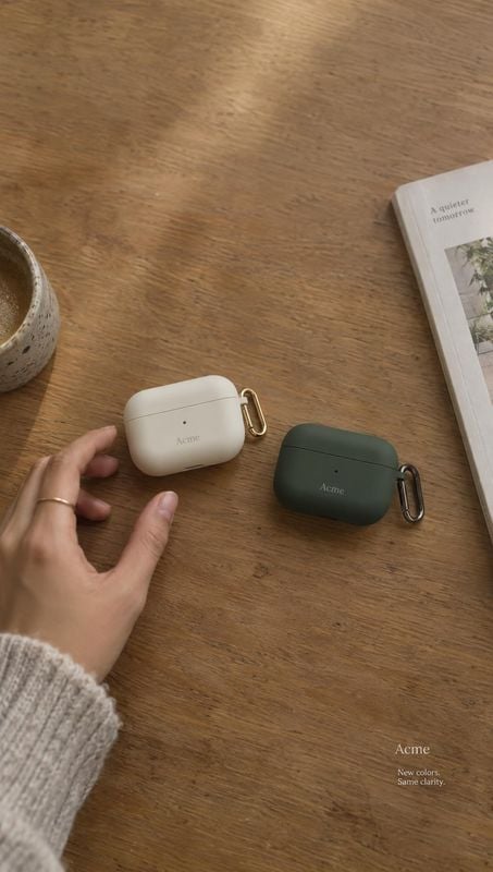 wireless earbud cases lifestyle scene product photo: Two neutral-toned wireless earbud cases with gold clips rest on a warm wooden table beside a hand, a ceram…