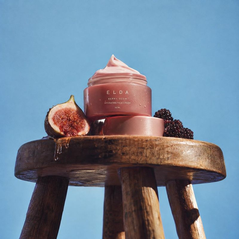 hydrating face cream ingredients product photo: A rose-toned face cream jar sits on a rustic wooden stool with a cut fig and blackberries against a clear blue…