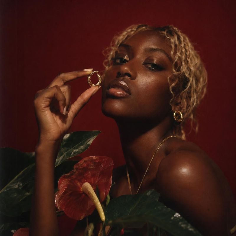 gold hoop earrings on-body detail product photo: A model with short blonde curls holds a gold hoop earring near her face, posed among glossy tropical leaves an…