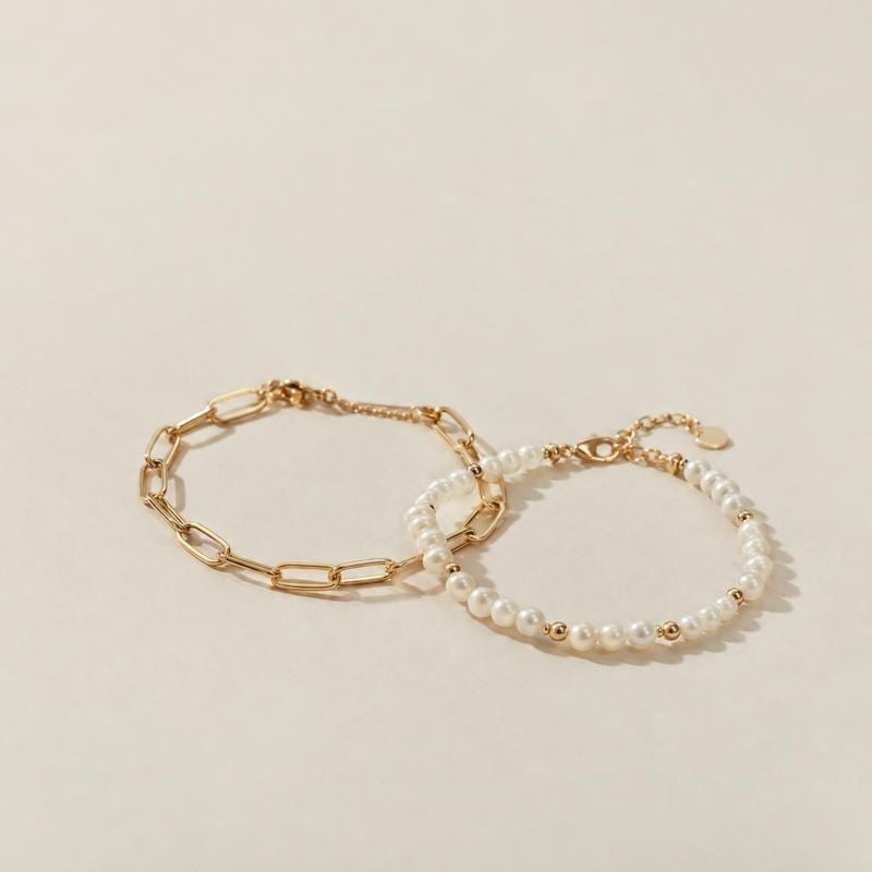 bracelets flat lay product photo: Two gold-tone bracelets, one with a linked chain and one with pearl-like beads, rest on a warm neutral surface in soft studio…
