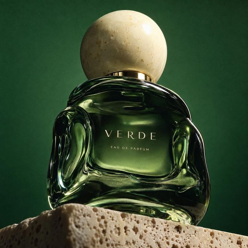 perfume beauty product photo: A sculptural green perfume bottle with a rounded stone-like cap rests on porous stone against a deep green backdrop, lit dramatic…