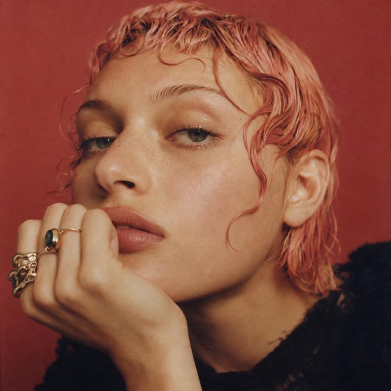 statement rings beauty close-up product photo: A close-up editorial portrait shows a woman with short pink hair resting her chin on a hand adorned with ornate…
