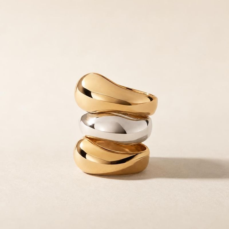 stacked rings jewelry product photo: Three sculptural gold and silver rings are stacked on a warm cream surface, softly lit with generous negative space and a…