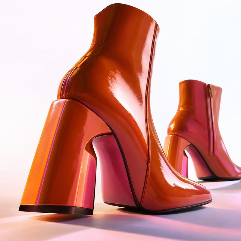 glossy orange ankle boots fashion product photo: Glossy orange-red ankle boots with sculptural block heels are arranged against a bright seamless studio backdr…