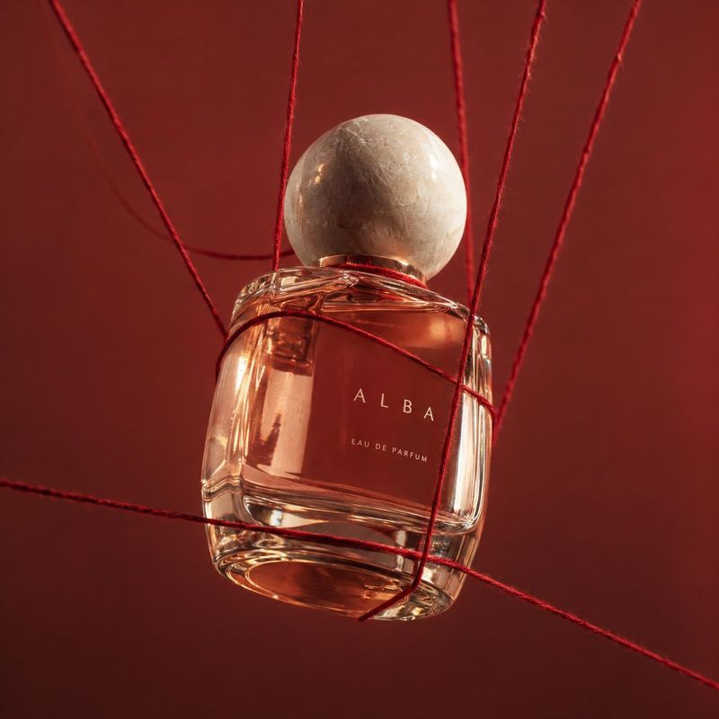 perfume floating product product photo: A glass perfume bottle with a pale spherical cap is suspended among taut red strings against a deep red backdrop, lit w…