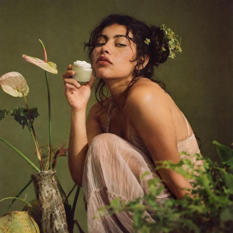 face cream beauty close-up product photo: A woman in a soft, sheer dress holds a small jar of cream among flowers and greenery against an olive studio backdrop…