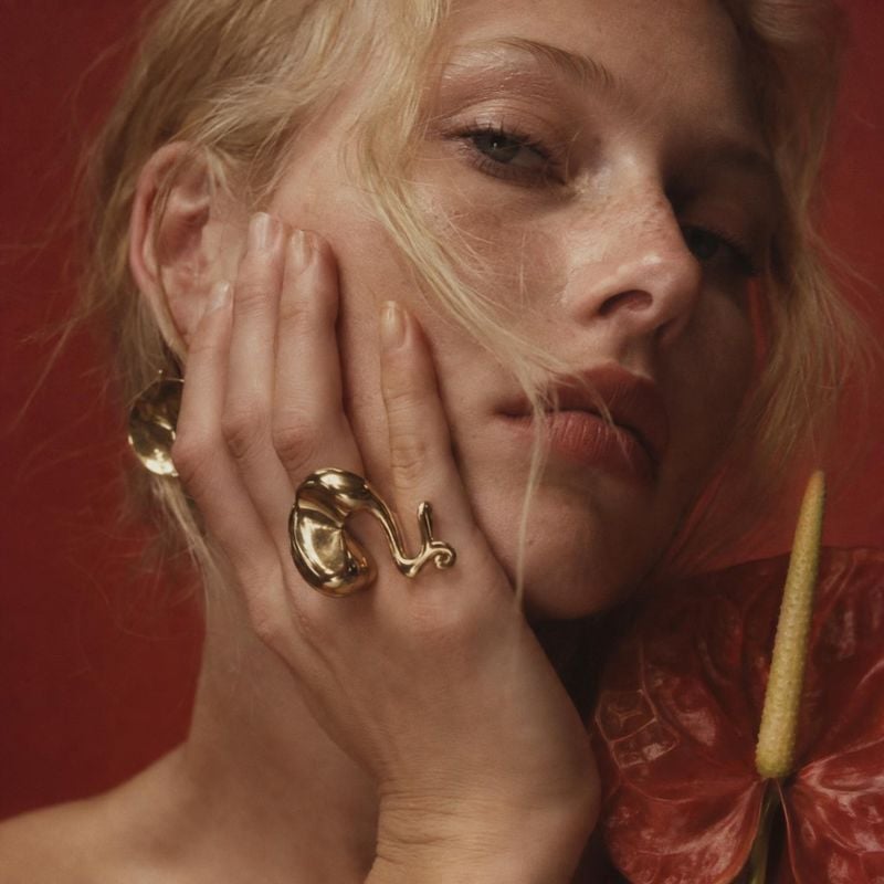 gold statement ring and earrings on-body detail product photo: A close fashion portrait shows a blonde model wearing sculptural gold jewelry against a deep red…